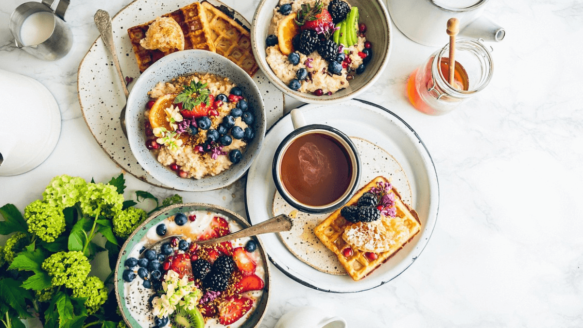 visão panorâmica de o que comer saudável para emagrecer em uma mesa branca com café da manha, tigelas de frutas, waffles, paes e iogurte. O que comer no café da manhã saudável para emagrecer